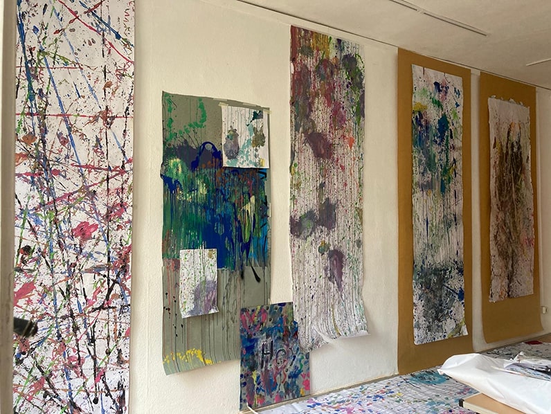 Abstract paintings with splattered and dripping paint on vertical canvases hanging on a white wall in an art studio.