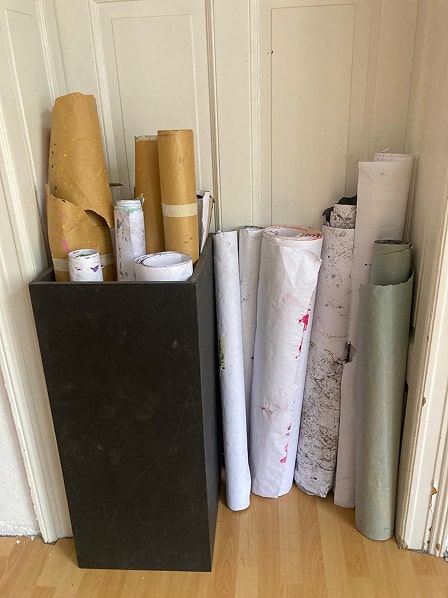 Several large rolls of paper and brown kraft paper stored upright in and next to a tall black rectangular container against a cream wall and wooden floor.