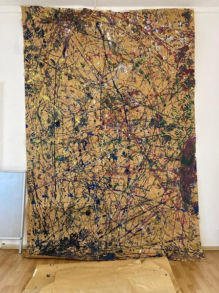 Large vertical abstract painting with chaotic multicolored splatters and drips on a beige background, displayed against a white wall over wooden floor.