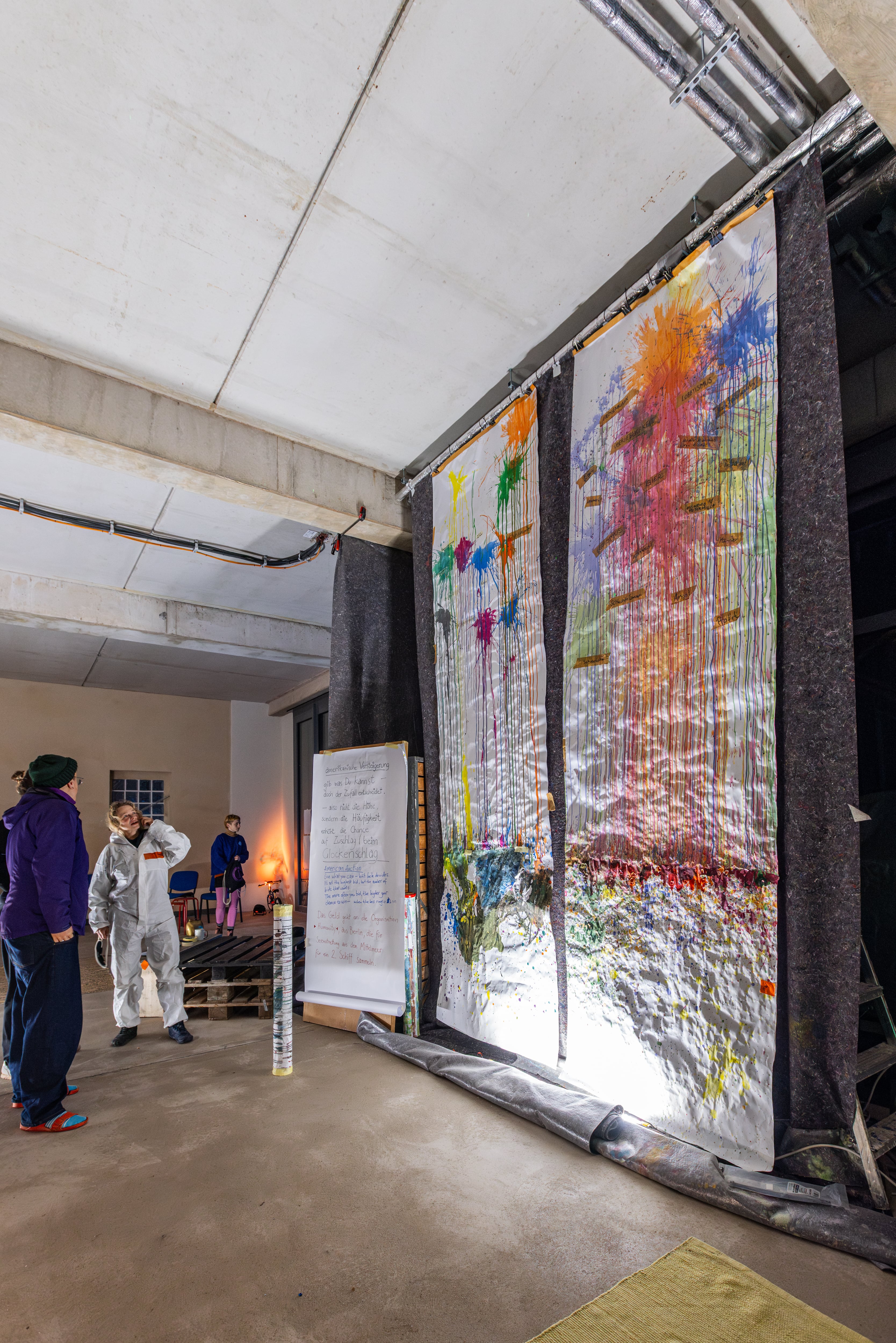 Indoor art space with large paint-splattered canvases hanging and two people observing nearby.