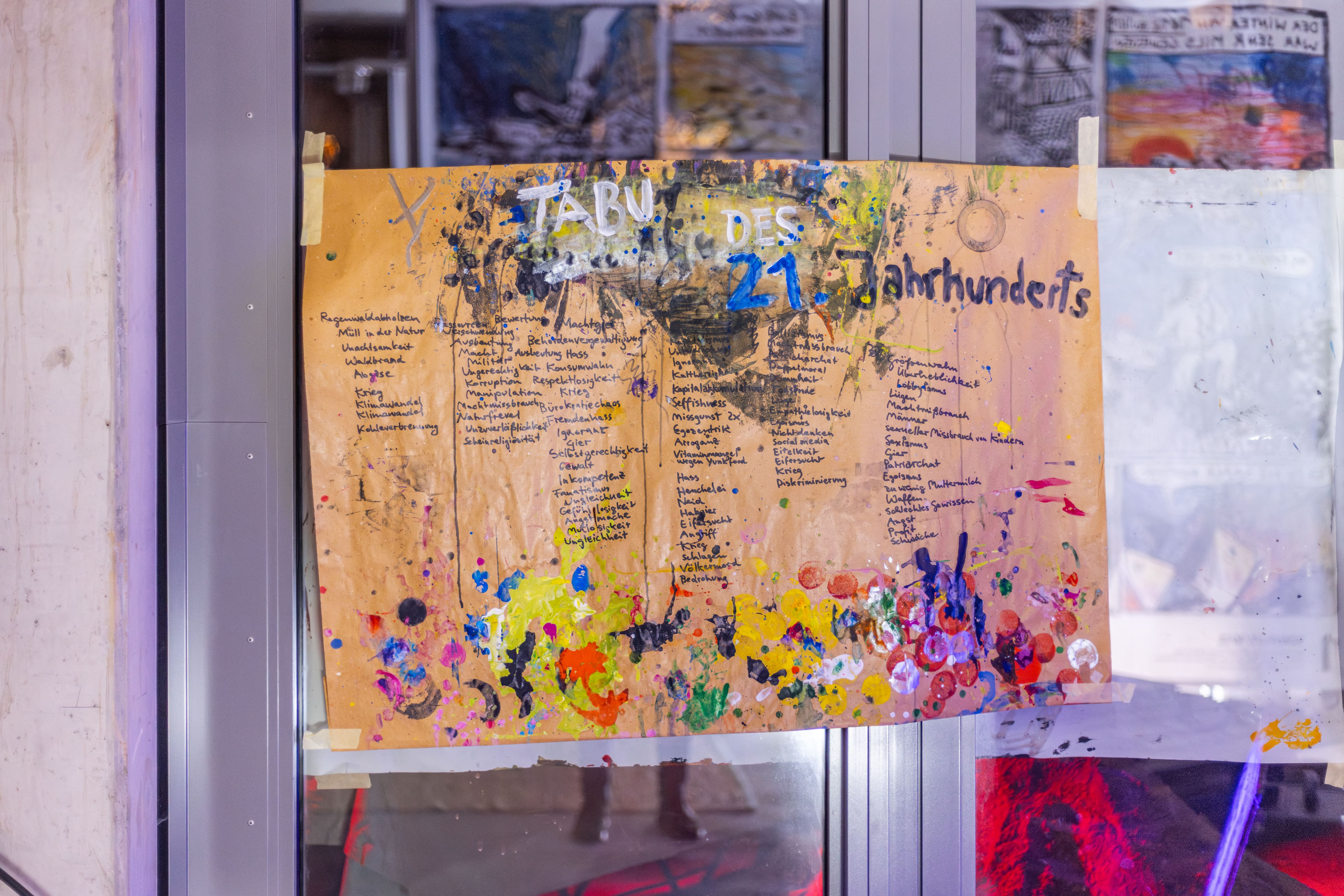 Handwritten German text titled 'Tabu des 21. Jahrhunderts' on brown paper taped to glass door with colorful paint splatters underneath.