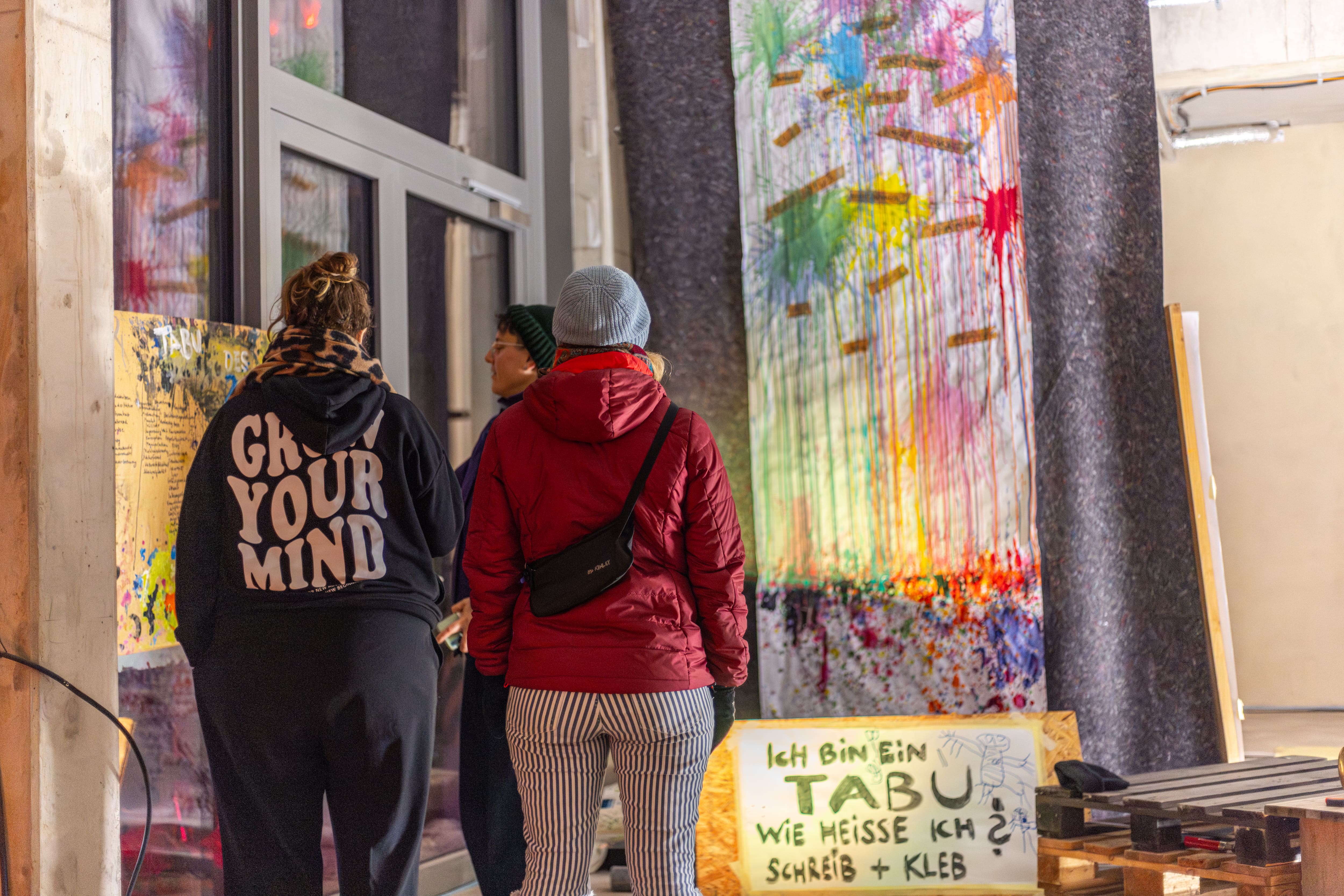 Three people from behind looking at colorful artwork inside a room, one wearing a black hoodie with 'GROW YOUR MIND' printed on the back.
