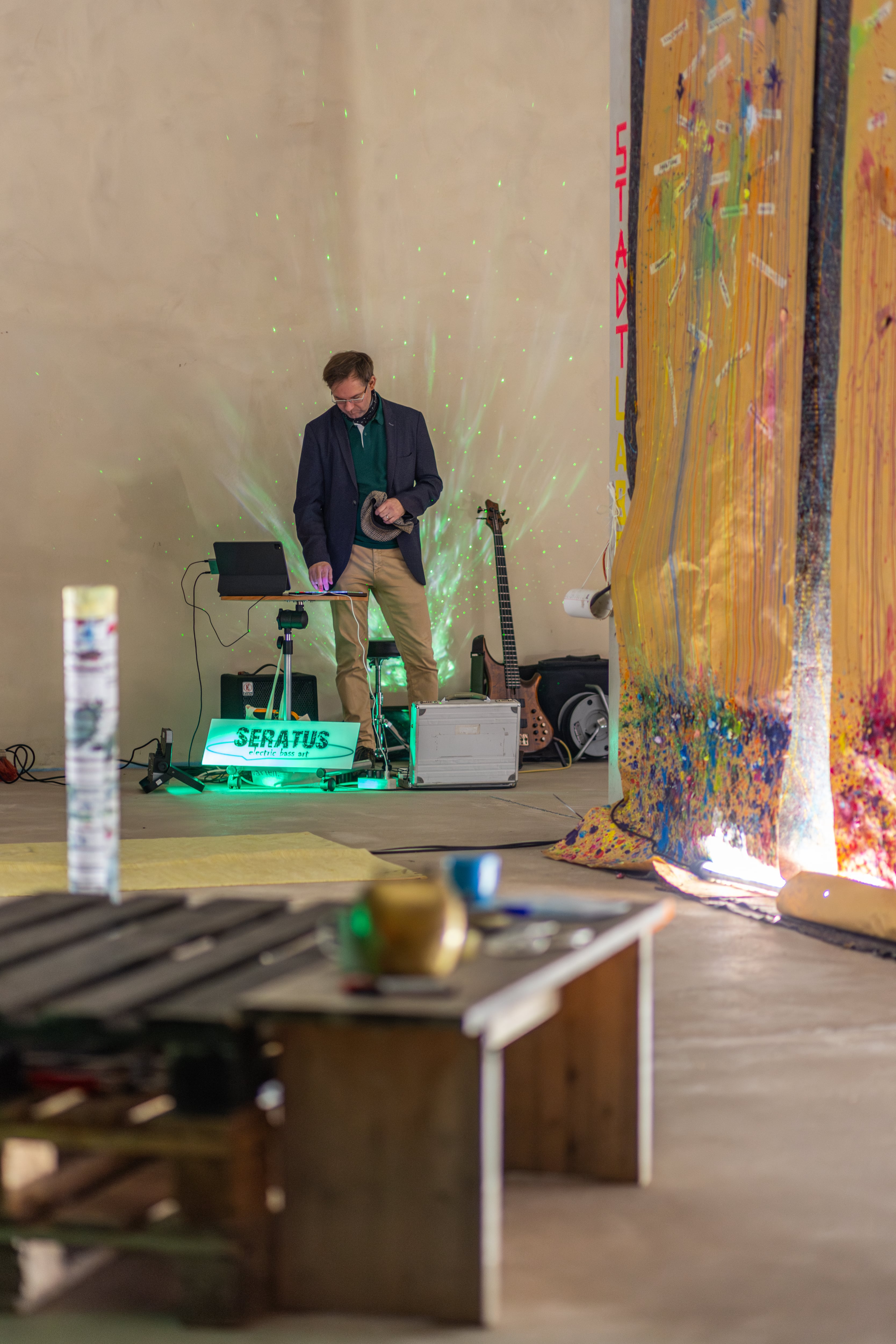 Man standing behind electronic musical equipment with green lighting and an electric bass guitar in the background in an indoor space.