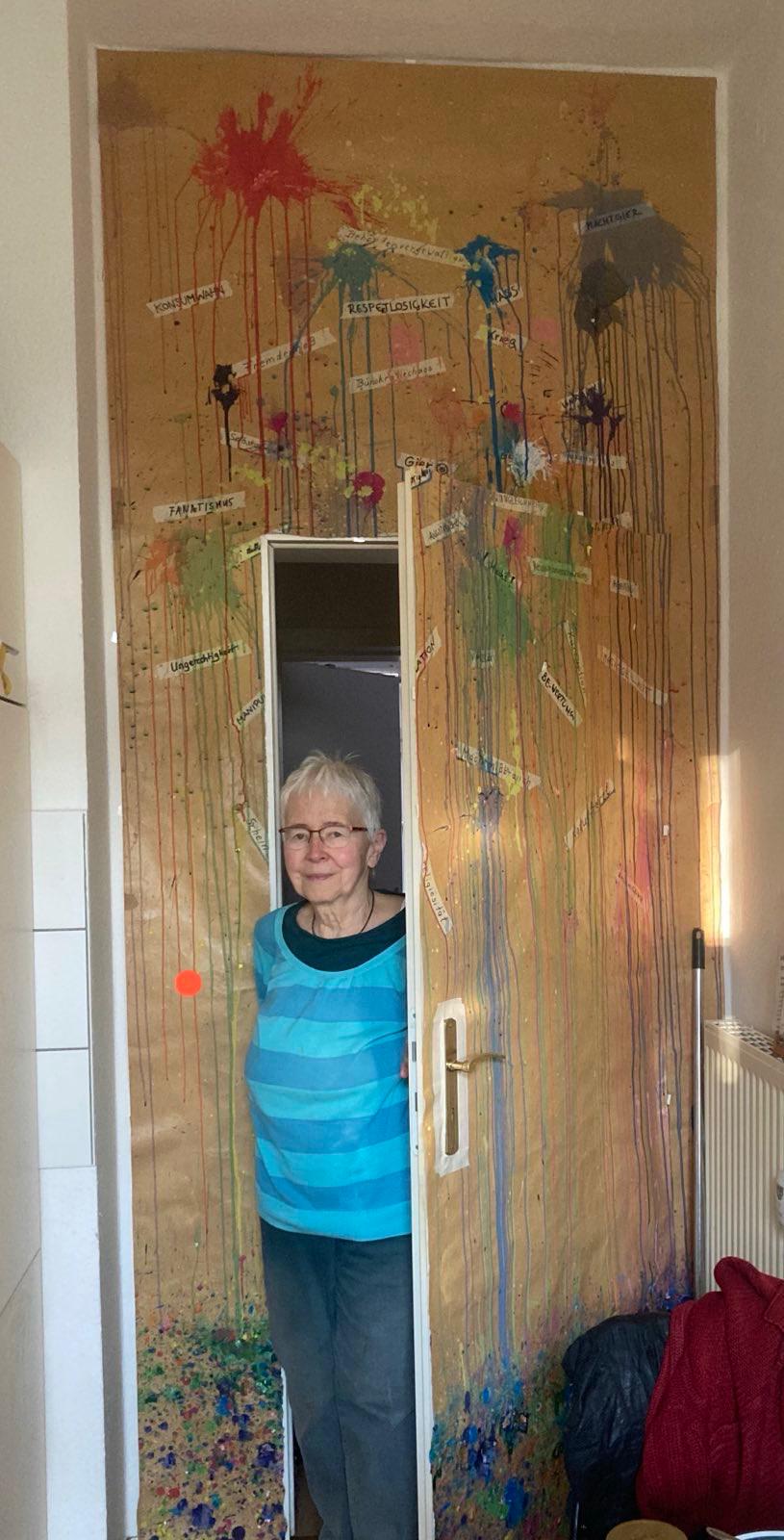 Older woman wearing glasses and a blue striped shirt standing in a doorway decorated with colorful paint splatters and German words on paper strips.