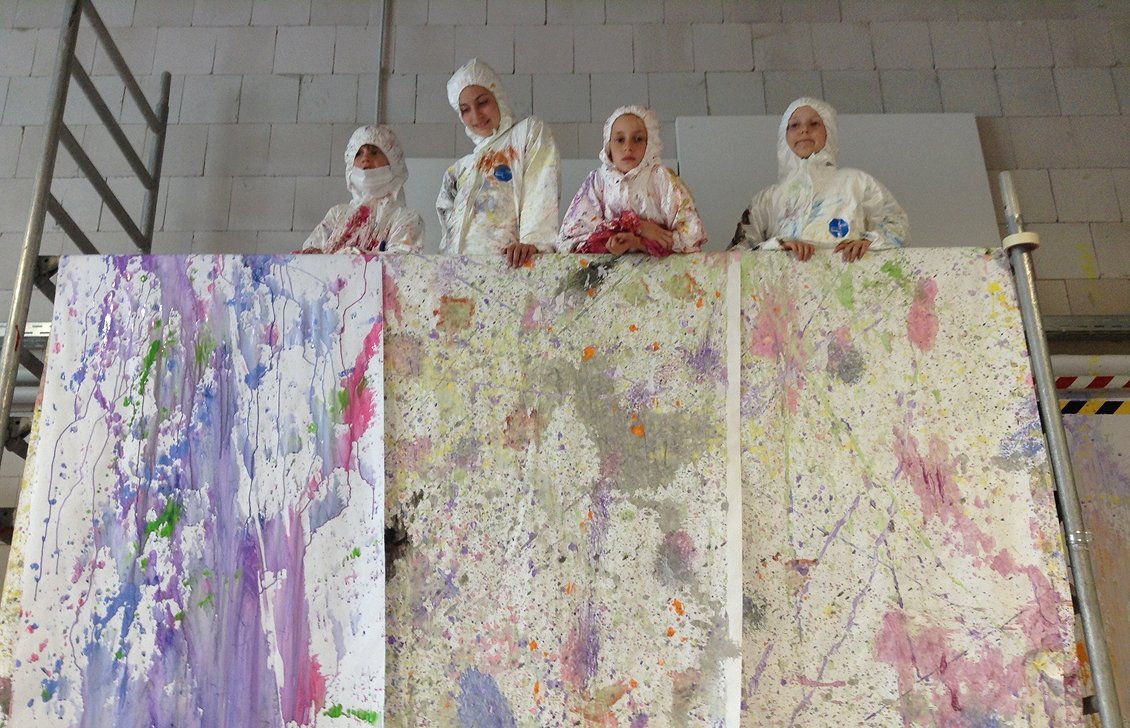 Four children in white paint-splattered protective suits looking over a large canvas covered in colorful paint splatters.