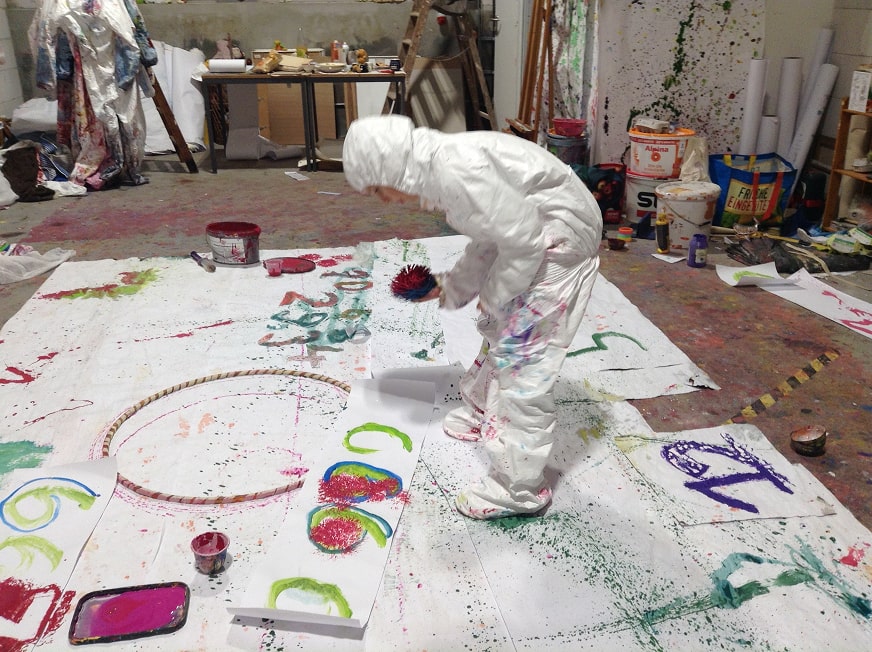 Person in white protective suit painting colorful numbers and shapes on large sheets spread on the floor in an art studio.