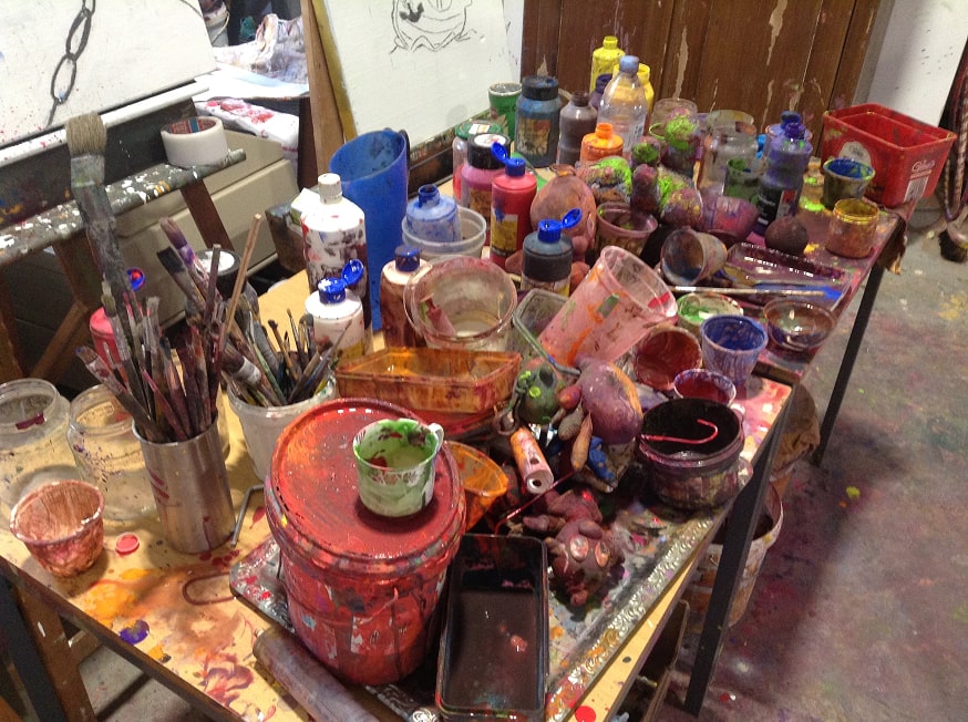 Cluttered artist workspace table covered with paintbrushes, paint bottles, cups, and containers stained with various bright paint colors.