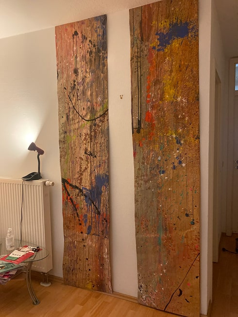 Two tall, narrow wooden panels leaning against a white wall, both covered in abstract multicolored paint splatters and drips in a room with a lamp, radiator, and glass table.