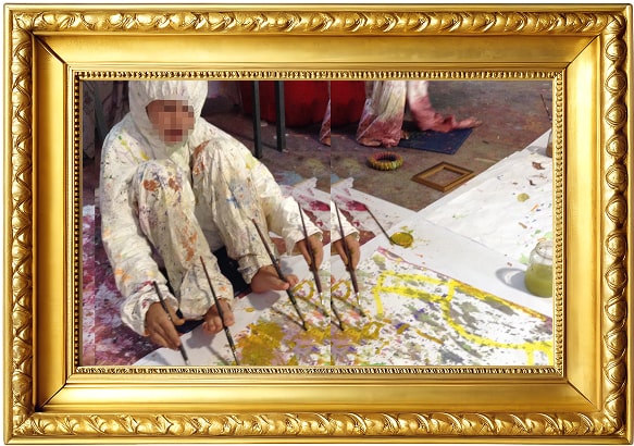 Person in white paint-splattered clothing painting on a large canvas on the floor using multiple brushes held by foot and hand.